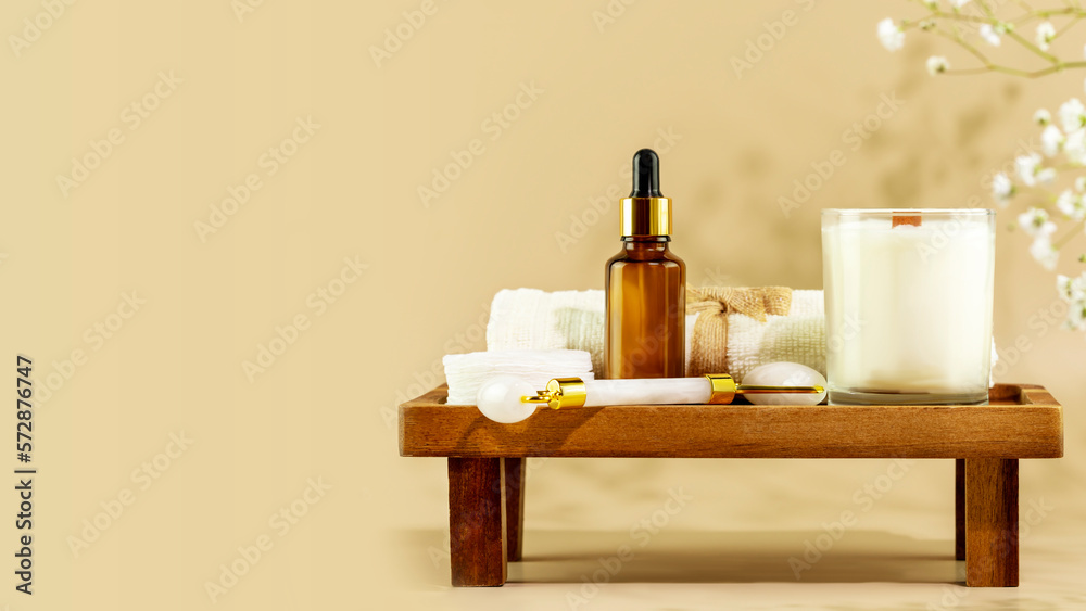 Eco friendly spa relax composition with serum, face roller, towel and candle on wooden podium on beige background with copy space. Wellness and skin care treatment. Concept of me time and self care Eco friendly spa relax composition with serum, face roller, towel and candle on wooden podium on beige background with copy space. Wellness and skin care treatment. Concept of me time and self care