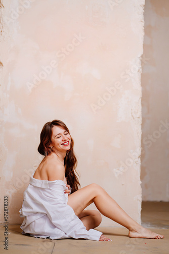 an attractive slender naked young woman in a white shirt sits on the floor. © andrey