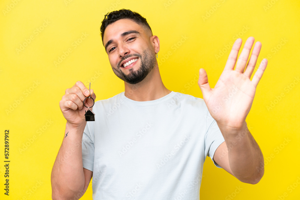 Young Arab man holding home keys isolated on yellow background saluting with hand with happy expression Young Arab man holding home keys isolated on yellow background saluting with hand with happy expression