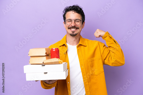 Young caucasian man holding fast food isolated on purple background doing strong gesture