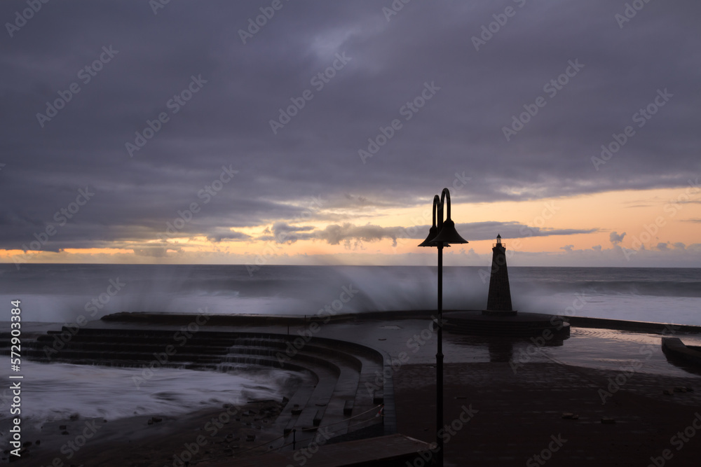 Sunset view of the small fishing pier of Bajamar, while the waves of the stormy sea break against the pier. Bajamar, Tenerife. Canary Islands. Spain