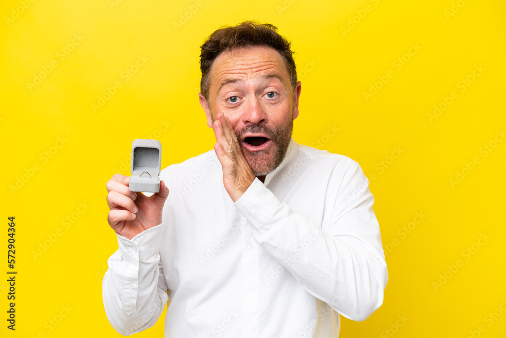 Middle age man holding a engagement ring isolated on yellow background whispering something Middle age man holding a engagement ring isolated on yellow background whispering something