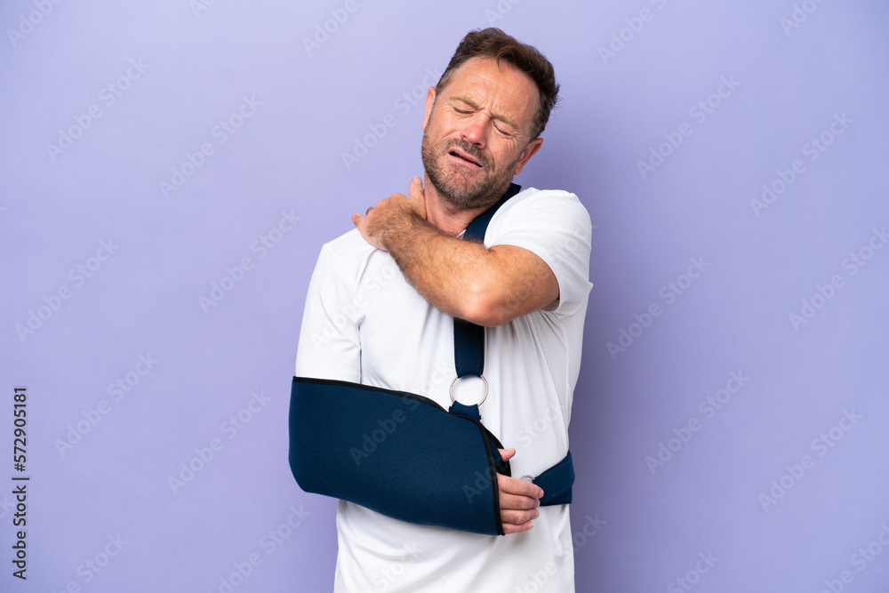Middle age caucasian man with broken arm and wearing a sling isolated on purple background suffering from pain in shoulder for having made an effort Middle age caucasian man with broken arm and wearing a sling isolated on purple background suffering from pain in shoulder for having made an effort