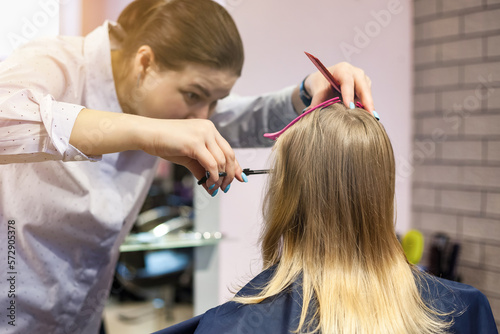 Barber woman makes hairdo for kid in barber shop. Hairdresser make fashionable hairstyle for little blond girl child in barbershop. Hairstyle, hair salon, beauty concept. Copy advertising text space