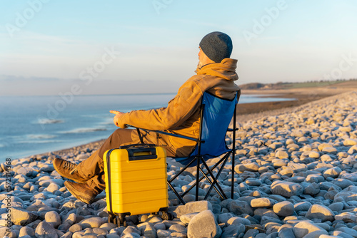Bearded Man in brown jacket with yellow suitcase relaxing alone and sitting on beach chairs on the seaside  at sunrise. Travel Lifestyle concept © Iryna