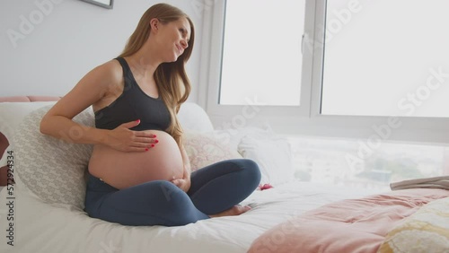 Pregnant woman relaxing sitting on bed at home wearing fitness clothing at home touching stomach and feeling baby kicking  - shot in slow motion