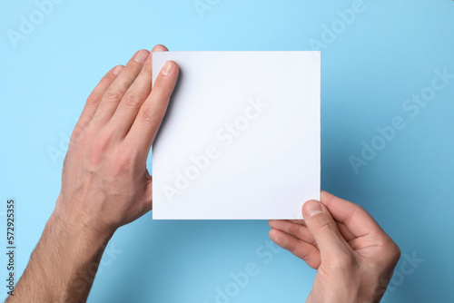 Man holding sheet of paper on light blue background, top view. Mockup for design