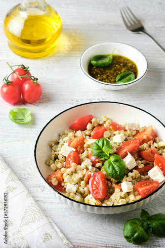 Warm salad of boiled pearl barley, ripe tomatoes, feta cheese with pesto sauce in a white plate on a light wooden background.