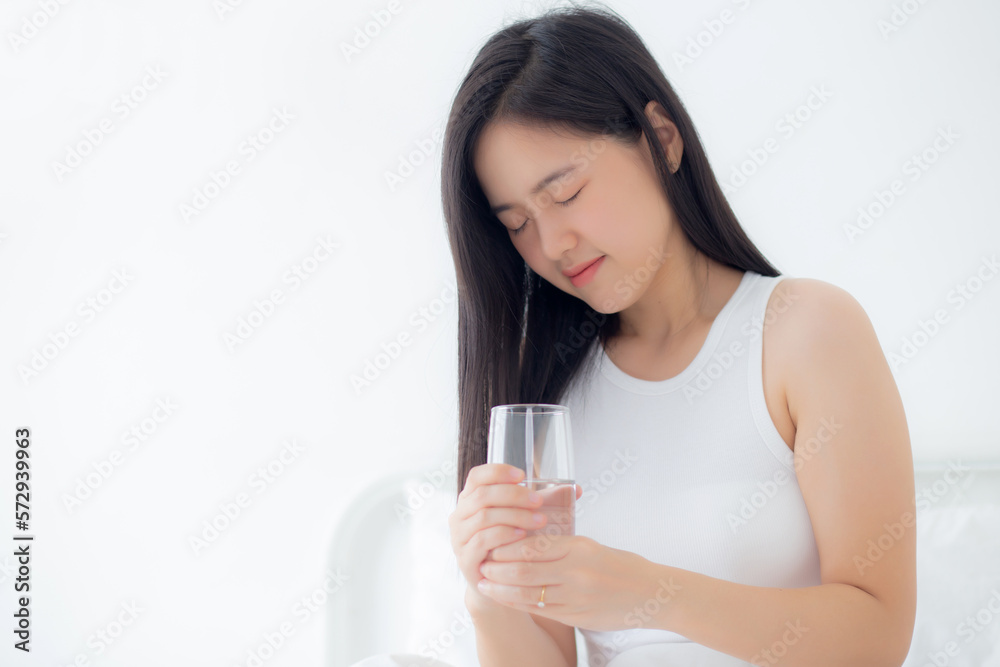 Beautiful young asian woman sitting on bed holding water of glass and drinking in the morning in bedroom at home, female drinking beverage mineral for healthy and nutrition, lifestyles concept. Beautiful young asian woman sitting on bed holding water of glass and drinking in the morning in bedroom at home, female drinking beverage mineral for healthy and nutrition, lifestyles concept.