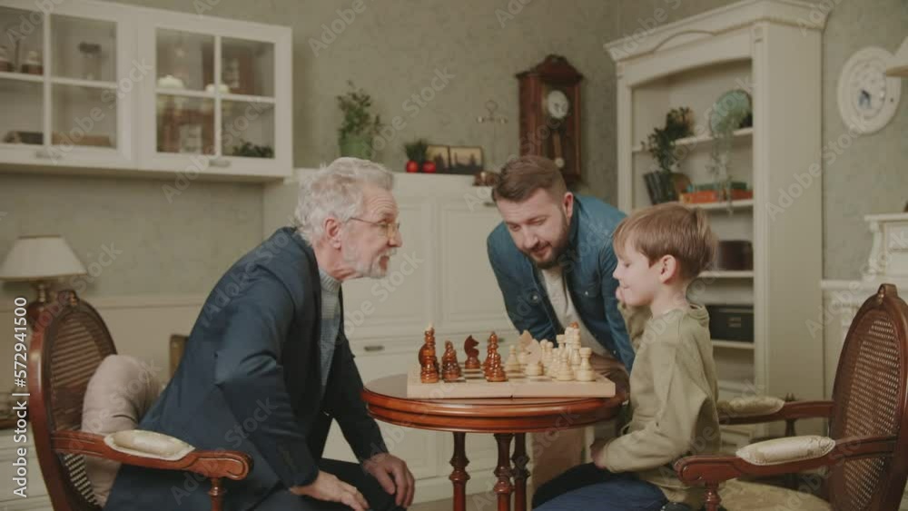 Three generations of men from the same family play chess at home. The grandson makes a move by rearranging a piece on the chessboard. Dad and Grandpa are watching. A family hobby. Three generations of men from the same family play chess at home. The grandson makes a move by rearranging a piece on the chessboard. Dad and Grandpa are watching. A family hobby.