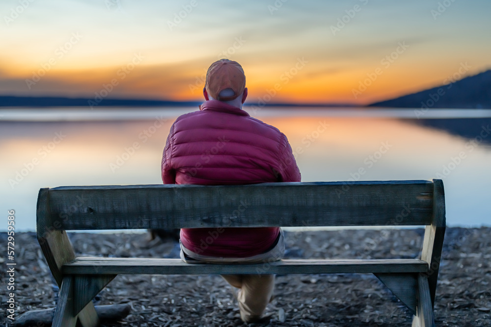 Φωτογραφία Winter scene of a man with red jacket in the middle of a bench looking out over a lake at sunset
