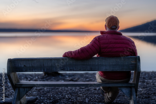 Winter scene of a man with red jacket on the right side of a bench looking out over a lake at sunset. Taking time for personal reflection, introspection, thinking about the past or the future. 