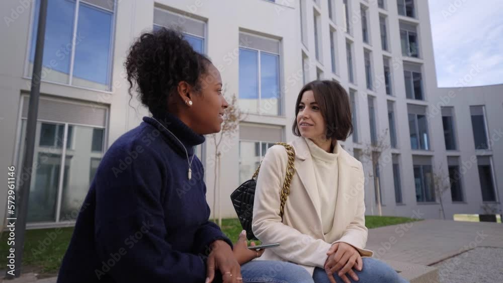 Two young female workers talking business and sitting outside a financial center. Couple of businesswomen coming to an agreement while taking a break. Meeting of empowered women entrepreneurs. Two young female workers talking business and sitting outside a financial center. Couple of businesswomen coming to an agreement while taking a break. Meeting of empowered women entrepreneurs.