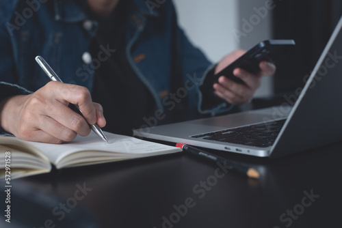 Business woman sitting at table using mobile phone, online working on laptop and making notes in notebook, calendar event planner, diary. Online education, e-learning concept