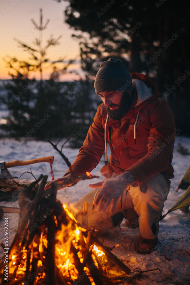 Fototapeta premium A man sits near a campfire, warming himself by the fire in a winter forest at sunset.