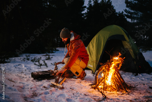 A tourist chops wood for a bonfire in a winter forest at night.
