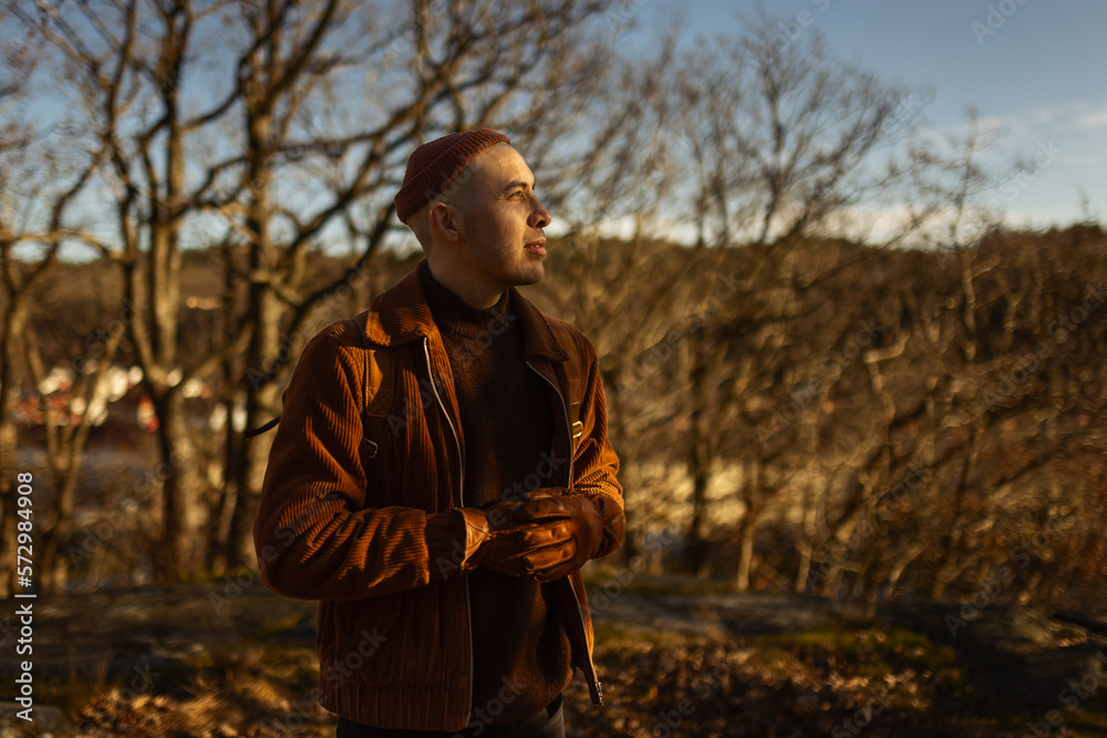 A caucasian man walking in the forest at the top of a hill at a lookout point wearing a leather backpack and a beanie.