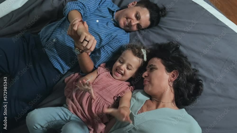 Top view of happy Caucasian lesbian couple and their beloved little daughter playing while lying on bed at home together Top view of happy Caucasian lesbian couple and their beloved little daughter playing while lying on bed at home together