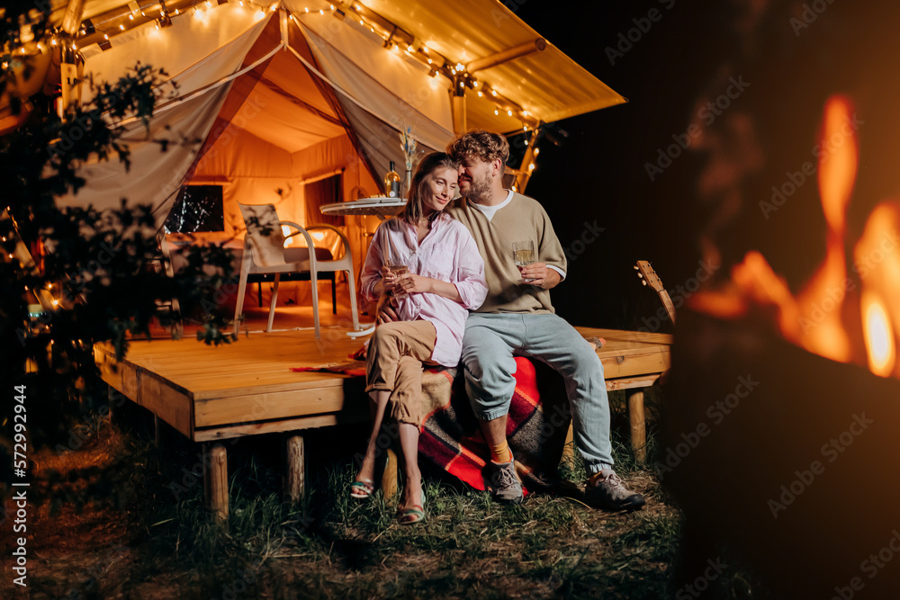 Happy couple relaxing in glamping on summer evening and drinking wine near cozy bonfire. Luxury camping tent for outdoor recreation and recreation. Lifestyle concept - obrazy, fototapety, plakaty 