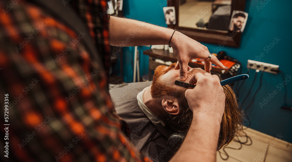 A professional barber cuts his beard to a young hipster man.