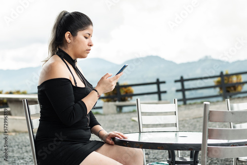 young latina woman, looking at her cell phone, while sitting in an outdoor café
