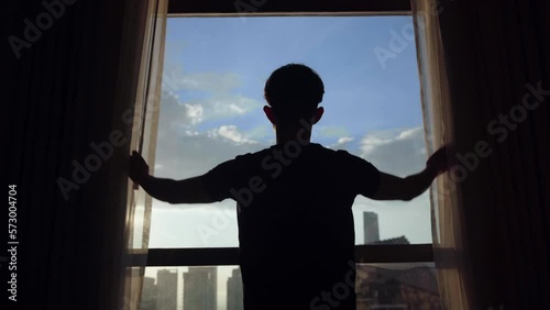 A young man opens the curtains in the morning, raises his arms and stretches. A cinematic man opens large window shades and let the sunlight into the room