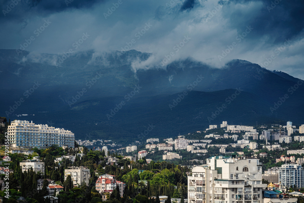 Panoramic view from height to city amidst thick trees against huge mountains with low clouds in daylight.