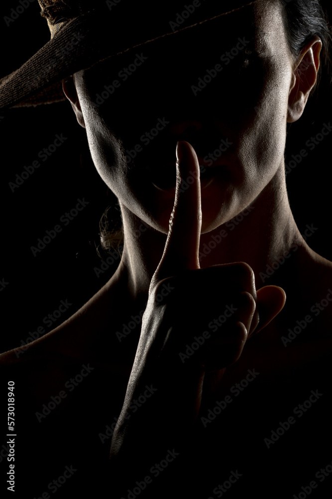 Silhouette of unknown woman with a hat, and the face in the shadow holding finger on her lips on a black background Silhouette of unknown woman with a hat, and the face in the shadow holding finger on her lips on a black background