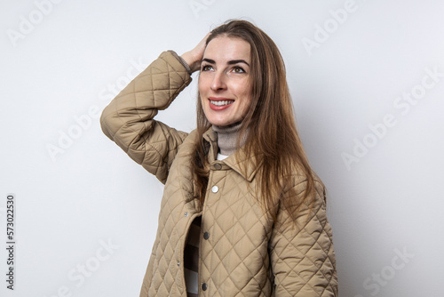 Smiling girl in a jacket straightens her hair near a white wall.