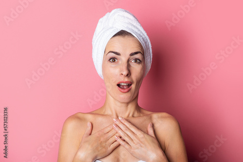 young woman with a towel on her head, bare shoulders, surprised, holds her hands on her chest on a pink background