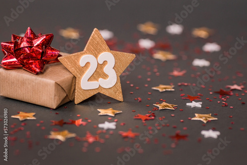 Greeting card for the Defender of the Fatherland Day. White, gold, red stars, a gift box and the number 23 on a brown background. The holiday is February 23.