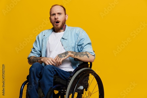 A man in a wheelchair surprised, with tattoos on his arms sits on a yellow studio background, the concept of health is a person with disabilities