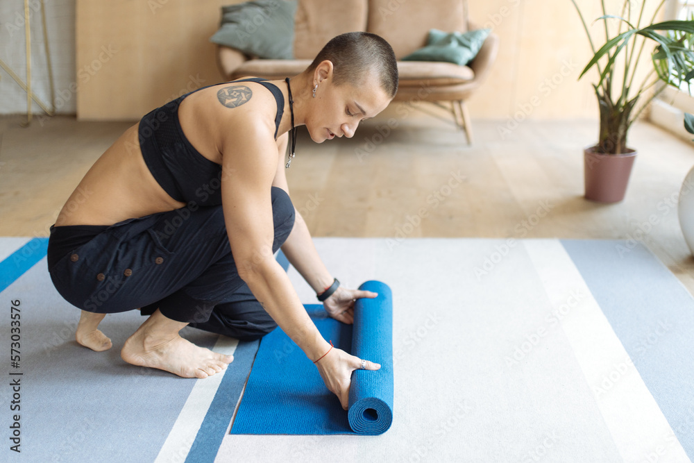 short haired tattooed woman unrolling yoga mat in home loft interior, preparing for yoga practice