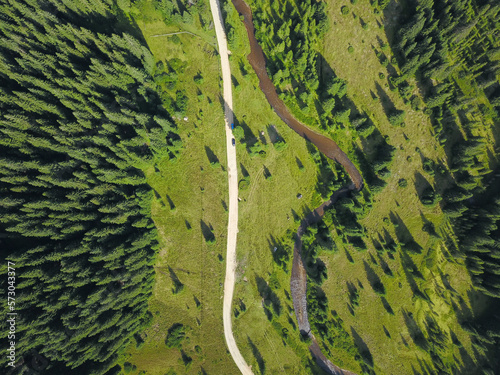 Aerial drone view of a forest gravel road winding along a green beautiful glade and spruce forests and a small stream. Sureanu Mountains, Romania. 