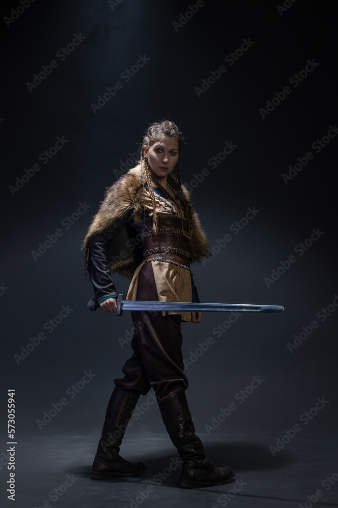 A young brave Viking heroine with a sword in hands on a dark background. A young woman in a suit in Scandinavian or fantasy style A young brave Viking heroine with a sword in hands on a dark background. A young woman in a suit in Scandinavian or fantasy style
