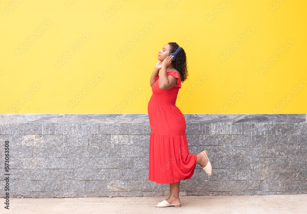Pregnant black woman walking on the sidewalk and listening streaming music on headphones with a yellow wall in the background.