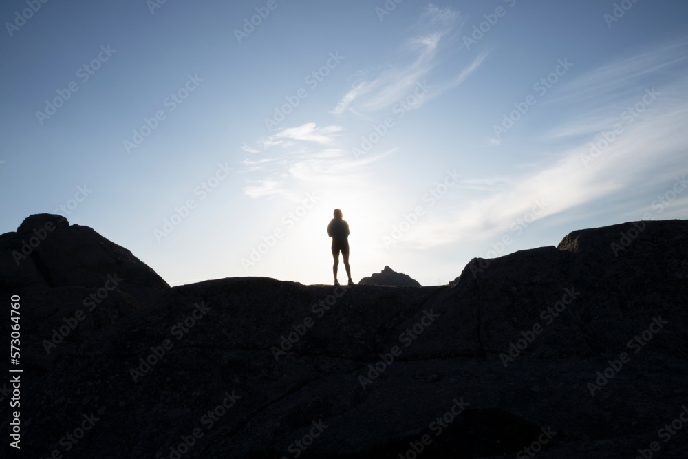 Hiking at sunset. Portrait of a young woman standing in the rocky hill with the sun hiding at her back. View of the female dark silhouette and sunny sky. 