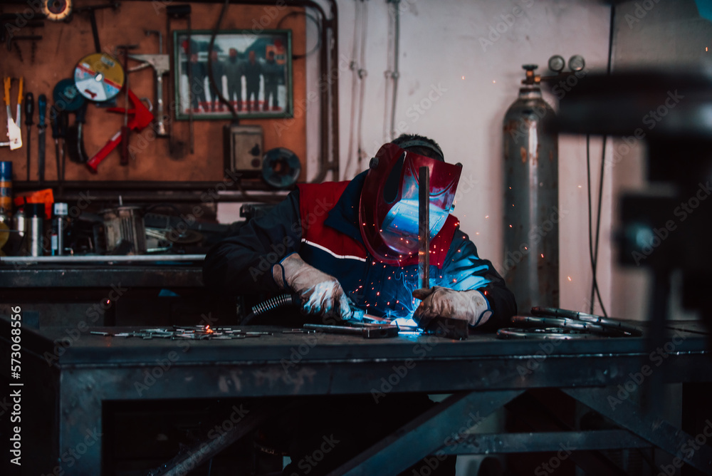 Close up welder in protective uniform and mask welding metal pipe on the industrial table with other workers behind in the industrial workshop. Close up welder in protective uniform and mask welding metal pipe on the industrial table with other workers behind in the industrial workshop.