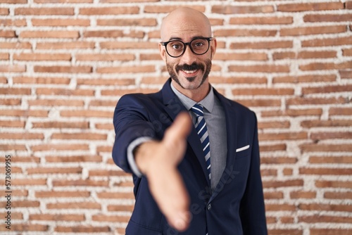 Bald man with beard wearing business clothes and glasses smiling friendly offering handshake as greeting and welcoming. successful business.