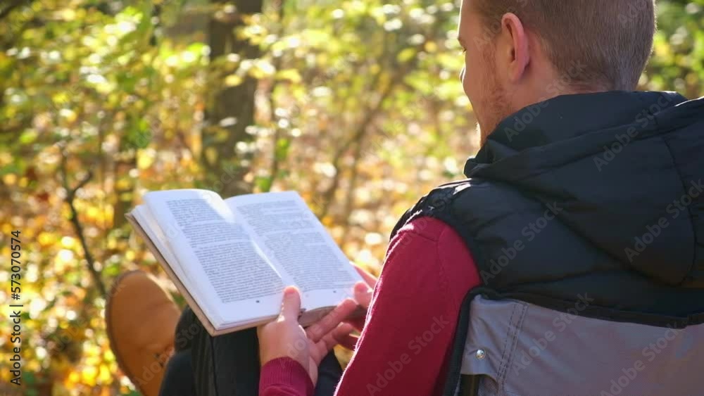 Young man is sitting on chair in an autumn park or forest and reading book. Mental recreation in nature. Quiet country life. Escapism. Digital detoxification. Concept of reading book in an autumn park