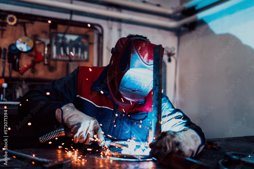 Profesional welder in protective uniform and mask welding metal pipe on the industrial table with other workers behind in the industrial workshop.