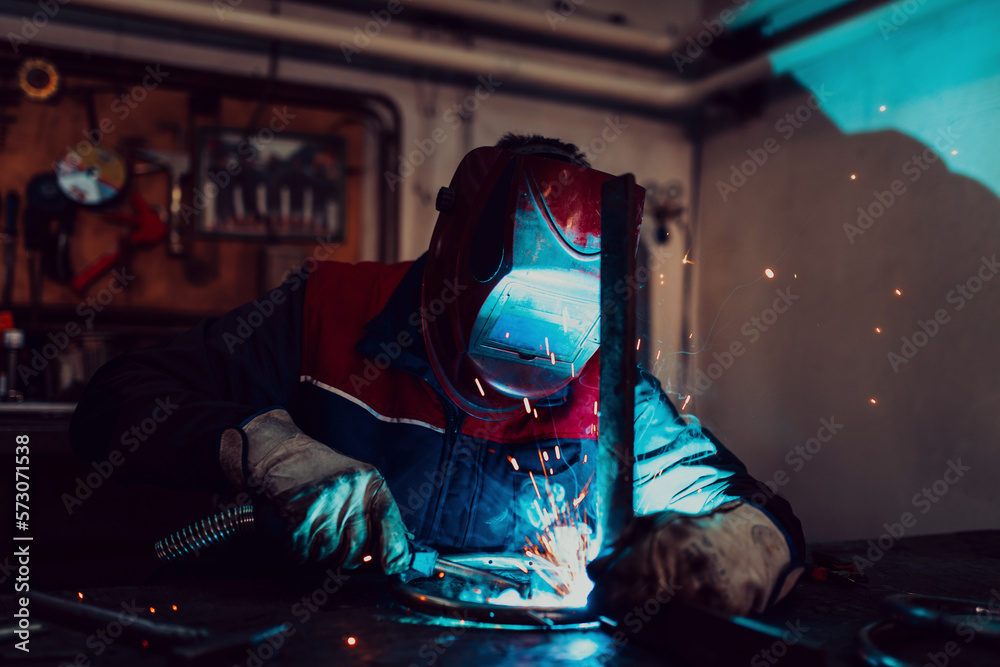 Profesional welder in protective uniform and mask welding metal pipe on the industrial table with other workers behind in the industrial workshop.