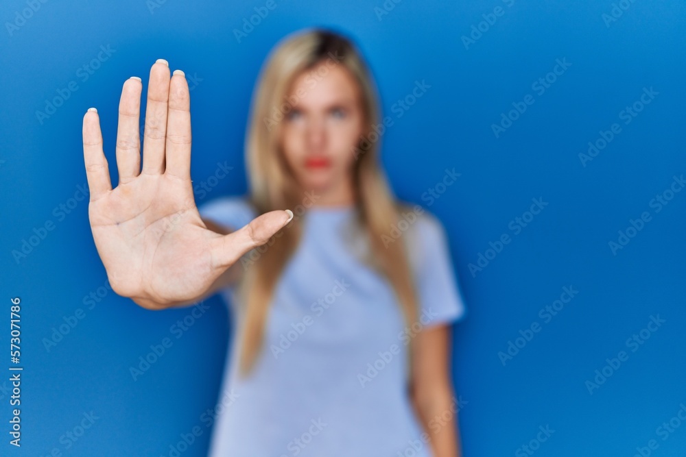 Beautiful blonde woman wearing casual t shirt over blue background doing stop sing with palm of the hand. warning expression with negative and serious gesture on the face.