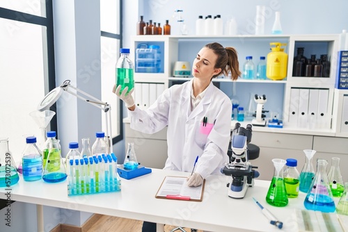 Young woman wearing scientist uniform holding bottle writing report at laboratory