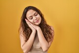 Young hispanic woman standing over yellow background sleeping tired dreaming and posing with hands together while smiling with closed eyes.