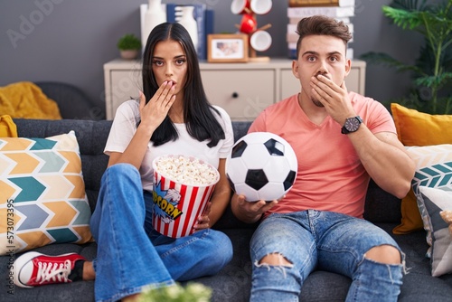 Young hispanic couple football hooligans holding ball and eating popcorn covering mouth with hand, shocked and afraid for mistake. surprised expression photo