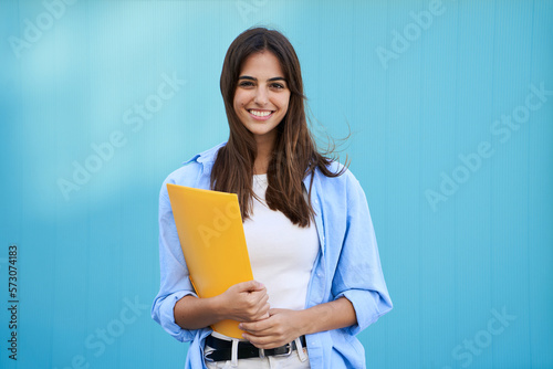Portrait on blue background of smiling and cheerful young Caucasian girl. Pretty student woman holding yellow folder. Happy college people posing and looking at camera. Copy space.