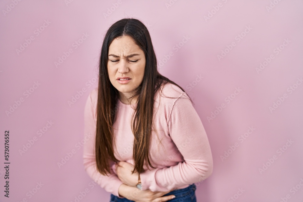 Fototapeta premium Young brunette woman standing over pink background with hand on stomach because indigestion, painful illness feeling unwell. ache concept.