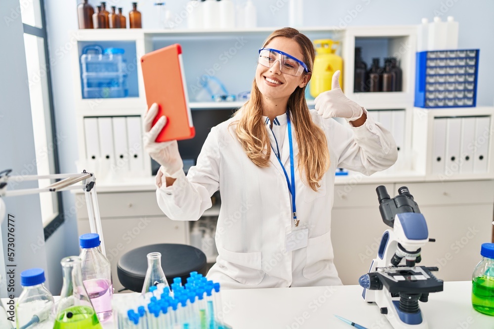 Young blonde scientist woman on video call working at laboratory smiling happy and positive, thumb up doing excellent and approval sign