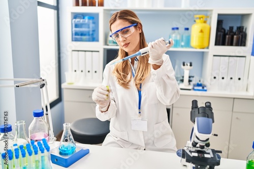Young blonde woman wearing scientist uniform pouring liquid using pipette at laboratory